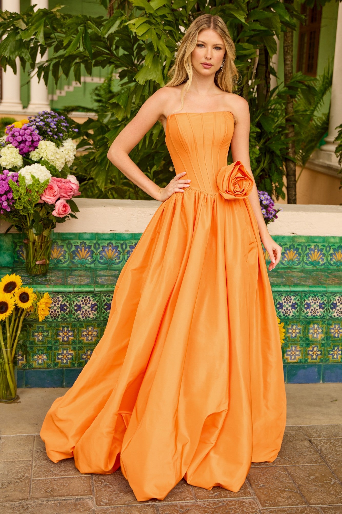 A woman stands outdoors before greenery, wearing the Ava Presley 47204 long prom dress featuring a strapless taffeta ball gown silhouette with a corset bodice and a large floral waist detail.